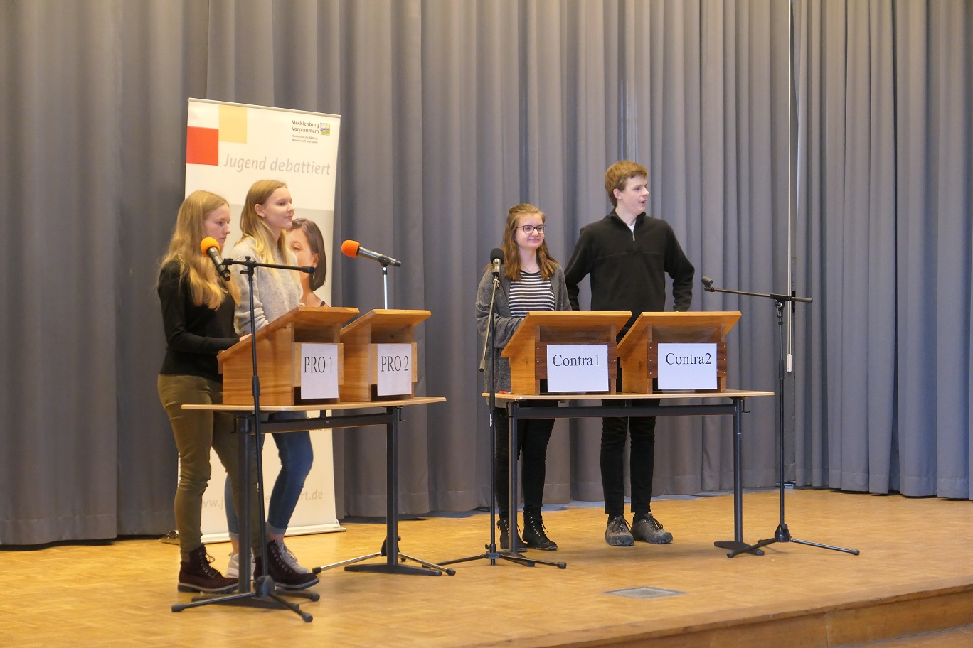 Vier Jugendliche diskutieren bei Jugend debattiert an Rednerpulten auf einer Bühne.